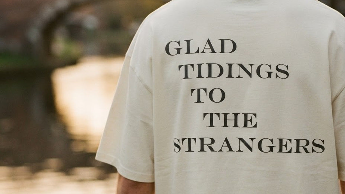 Back view of a cream ALUNA PATIENCE oversized t-shirt by a canal at golden hour, the back print reading Glad tidings to the strangers