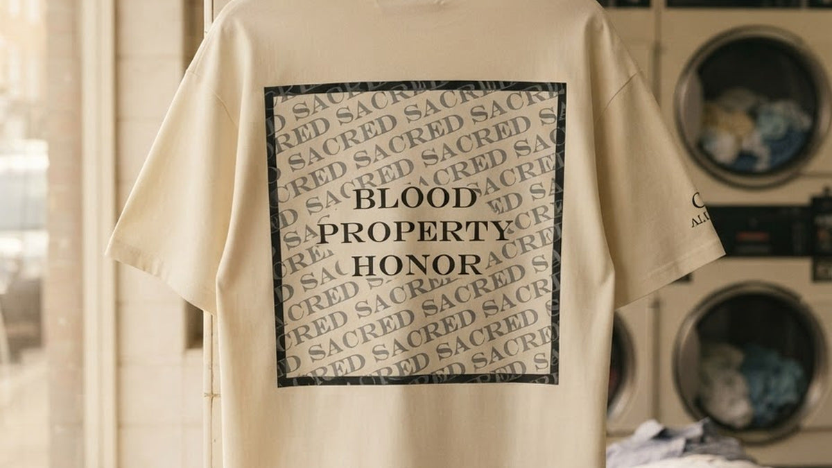 Cream ALUNA SACRED oversized t-shirt on a wooden hanger in a laundrette, back print reading BLOOD PROPERTY HONOR inside a square of repeated SACRED text